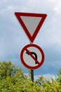 Give way and no left turn red and white signs mounted on strong metal pole surrounded with dense leaves on cloudy stormy blue sky