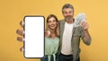 Giveaway concept. Emotional middle aged couple holding lot of dollar cash, showing smartphone with blank screen, mockup Royalty Free Stock Photo
