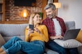 Glad adult caucasian female show phone to man with laptop, watch video in living room interior