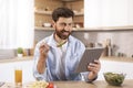 Glad adult caucasian man with beard eats lunch, read news at tablet, watch video in modern kitchen interior