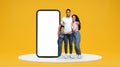 Glad arab millennial parents and small daughter hugs on yellow background with big smartphone