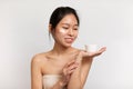 Glad asian lady with perfect skin applying moisturizing cream on face and holding jar with blank space, white background