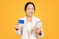 Glad Asian millennial woman in business suit, holds credit card, use smartphone for online shopping