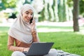 Glad attractive young middle eastern female in hijab looks in laptop, call by phone, works remotely in park