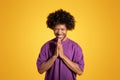 Glad black middle aged curly man in violet t-shirt prays with hands, isolated on orange background, studio