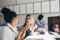 Glad blonde office worker posing with smile during brainstorm with colleagues. African female student in white shirt