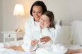 Glad caucasian little girl and young lady in bathrobe sit on white bed with headphones and smartphone