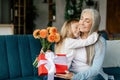 Glad caucasian little granddaughter hugging aged grandma with bouquet of flowers and box gift Royalty Free Stock Photo