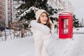 Glad caucasian woman in knitted hat goes to red phone booth in winter day. Outdoor portrait of attractive european girl Royalty Free Stock Photo