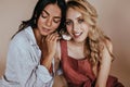 Glad curly blonde girl posing with tanned sister. Indoor photo of lovely latin female model sitting beside fair-haired Royalty Free Stock Photo