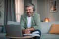 Glad european aged man retired with beard in headphones looks at laptop, work in room interior Royalty Free Stock Photo