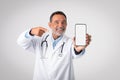 Glad european mature doctor in white coat with stethoscope points finger at smartphone