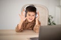 Glad european small kid in headphones waving hand, has video call with teacher, tutor at table