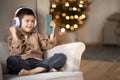 Glad european small kid in wireless headphones calls by phone, sits in armchair in living room interior