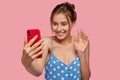 Glad European woman with pleasant smile, waves with hand at camera of cell phone, dressed in polka dot dress, models Royalty Free Stock Photo