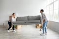 Glad european young male and female carry sofa in empty room with cardboard boxes