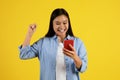 Glad excited young chinese lady looking at smartphone and making victory gesture with hand