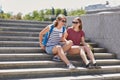 Glad female best friends wear shades, shorts and sneakers, carry rucksack, sit on stairs, return from university, enjoy sunny