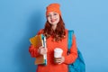 Glad ginger female student has coffee break after lectures, enjoys hot beverage, has rucksack on back, lady with wavy hair looks Royalty Free Stock Photo