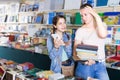 glad girl chatting on screen tablet and choosing literature books with mother