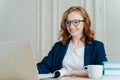 Glad lovely woman has ginger hair, positive smile, sits with laptop computer at desktop, happy to make video call or conference,