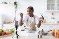 Glad millennial african american female in apron cooks at home and tasting dish shooting video lesson by phone Royalty Free Stock Photo