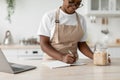 Glad millennial african american woman in glasses and apron writes new recipe with laptop in modern scandinavian kitchen Royalty Free Stock Photo