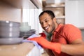 Glad millennial black man in rubber gloves wipes dust from shelf with dishes enjoy purely in white kitchen