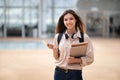 Glad millennial caucasian female brunette student with backpack, books and smartphone in wireless headphones