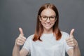 Glad positive woman with tender smile on face, has brown hair, raises two thumbs, demonstrates her approval, over grey Royalty Free Stock Photo