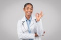 Glad pretty adult african american woman doctor in white coat make ok sign with hand