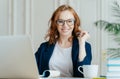 Glad redhead curly woman works freelance, uses laptop computer, writes down notes in notepad, drinks coffee, wears optical glasses