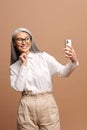 Glad senior woman is using smartphone for video connection isolated on beige, carefree mature gray-haired lady has Royalty Free Stock Photo