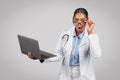Glad shocked pretty millennial black lady doctor in white coat with laptop, with open mouth, take off glasses