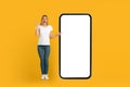 Glad shocked young european woman with open mouth in white t-shirt points finger at huge phone with blank screen