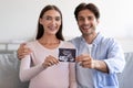 Glad smiling millennial european man and woman holding ultrasound photo of baby in living room