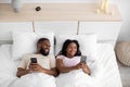 Glad smiling young black female and male, lying on bed, typing on phones, chatting, playing online game