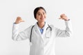 Glad smiling young indian female doctor in coat points fingers at herself, isolated on white background, studio Royalty Free Stock Photo