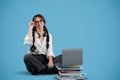 Glad teen girl with pigtails in glasses sits on floor with books, computer looks at copy space on blue background