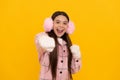 glad teen girl in winter mittens and earmuffs punching on yellow background, happiness Royalty Free Stock Photo