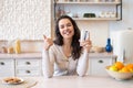 Glad woman holding glass with clean fresh water and showing thumb up, sitting in kitchen interior and smiling at camera Royalty Free Stock Photo