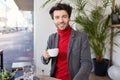 Glad young attractive unshaved brunette guy with trendy haircut holding cup of tea in raised hand and looking positively at camera