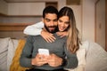 Glad young caucasian lady hugs arabic male watch video on smartphone, reads message, blog in living room