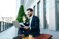 Glad young european businessman with beard in suit has video call on laptop, waving hand