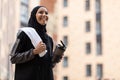 Glad young muslim lady in hijab and sportswear with phone on shoulder with cocktail cup and towel at street