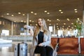 Happy pretty woman sitting in airport waiting room with tablet and grey valise. Royalty Free Stock Photo