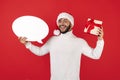 Gladful hipster man wears santa claus hat holding gift box and speech bubble over red background.