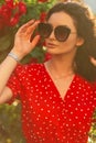 Glamorous young stylish curly girl with retro sunglasses in a trendy red bright dress in a summer garden on the background of a Royalty Free Stock Photo