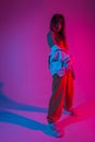 Glamorous young woman dancer in trendy sports youth clothes posing in a room with multi-colored pink-blue color. Attractive girl