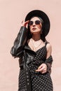 Glamorous young woman with fashion bag in stylish sunglasses in leather jacket in beautiful dress straightens hat on sunny day. Royalty Free Stock Photo